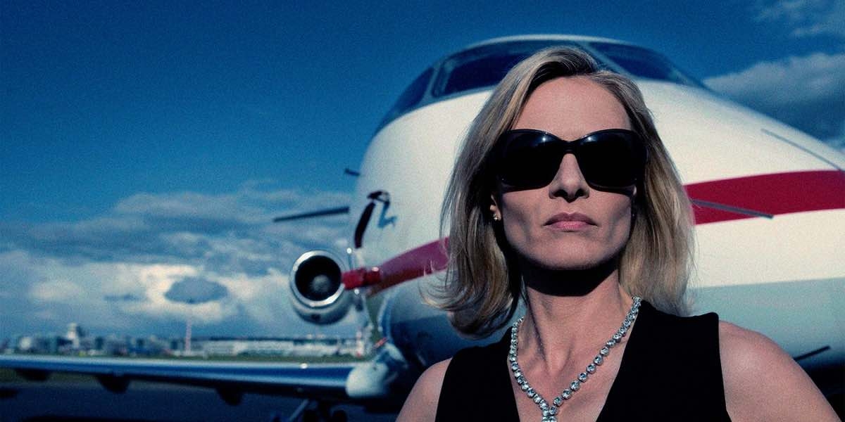 Rich blond woman in sunglasses and diamond necklace in front of a private jet.