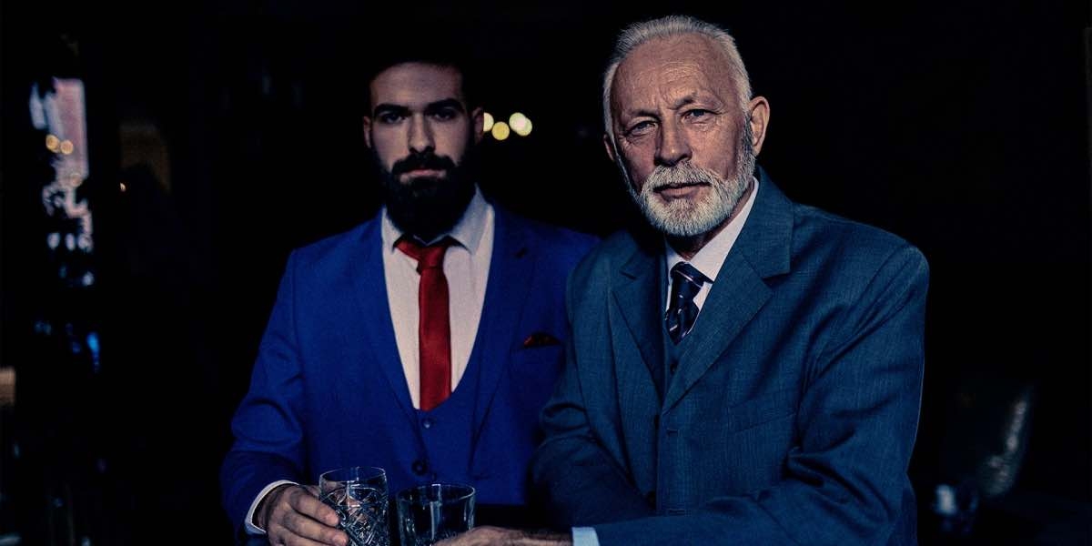 Two gentlemen in suits and ties looking distinguished, drinking at a bar, looking at the viewer.